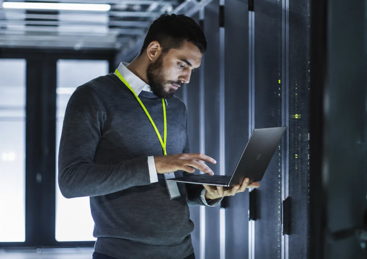 An engineer with a laptop in a server room, checking what is running.